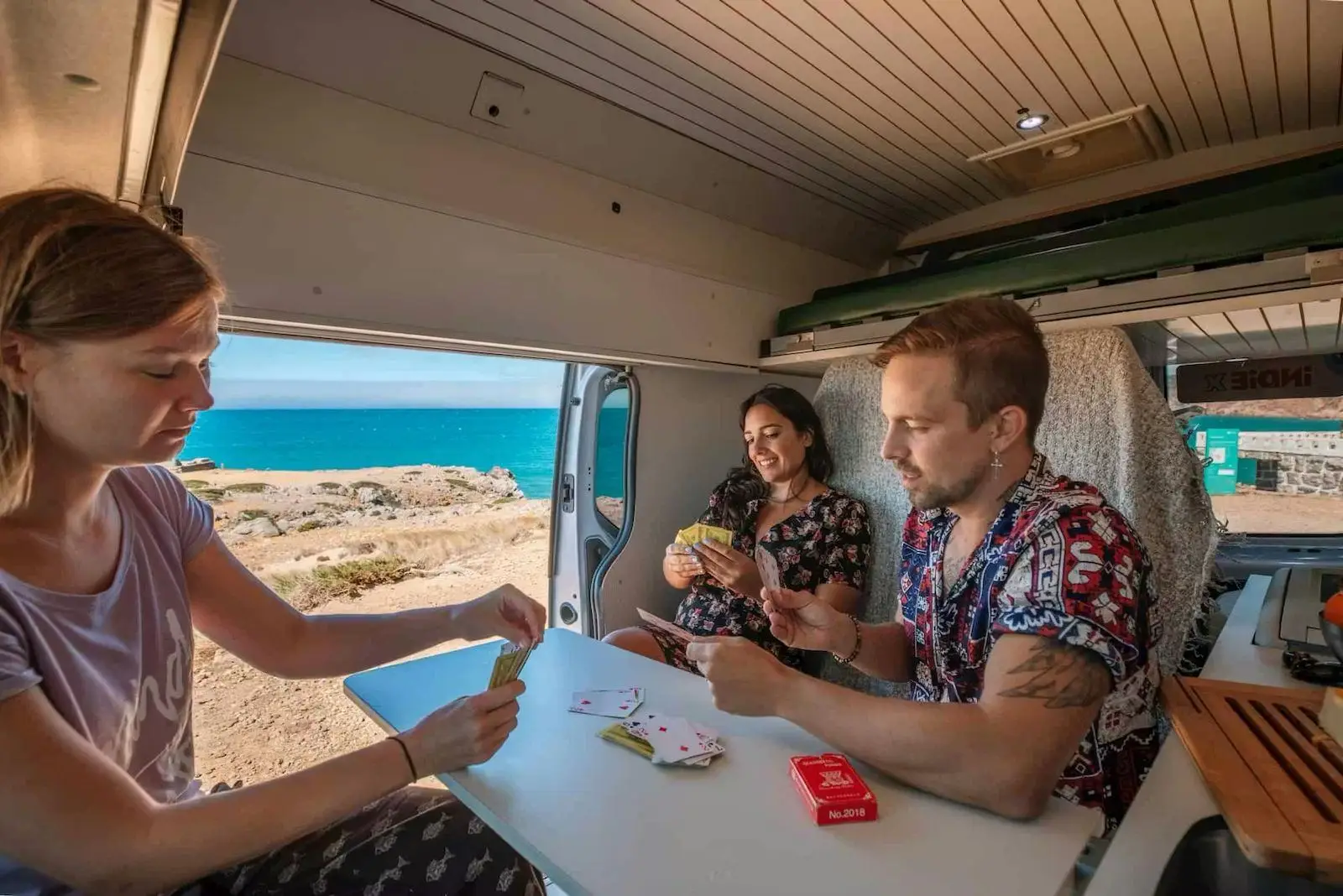Three people playing cards at the table in the Fiat Talento campervan by the coast