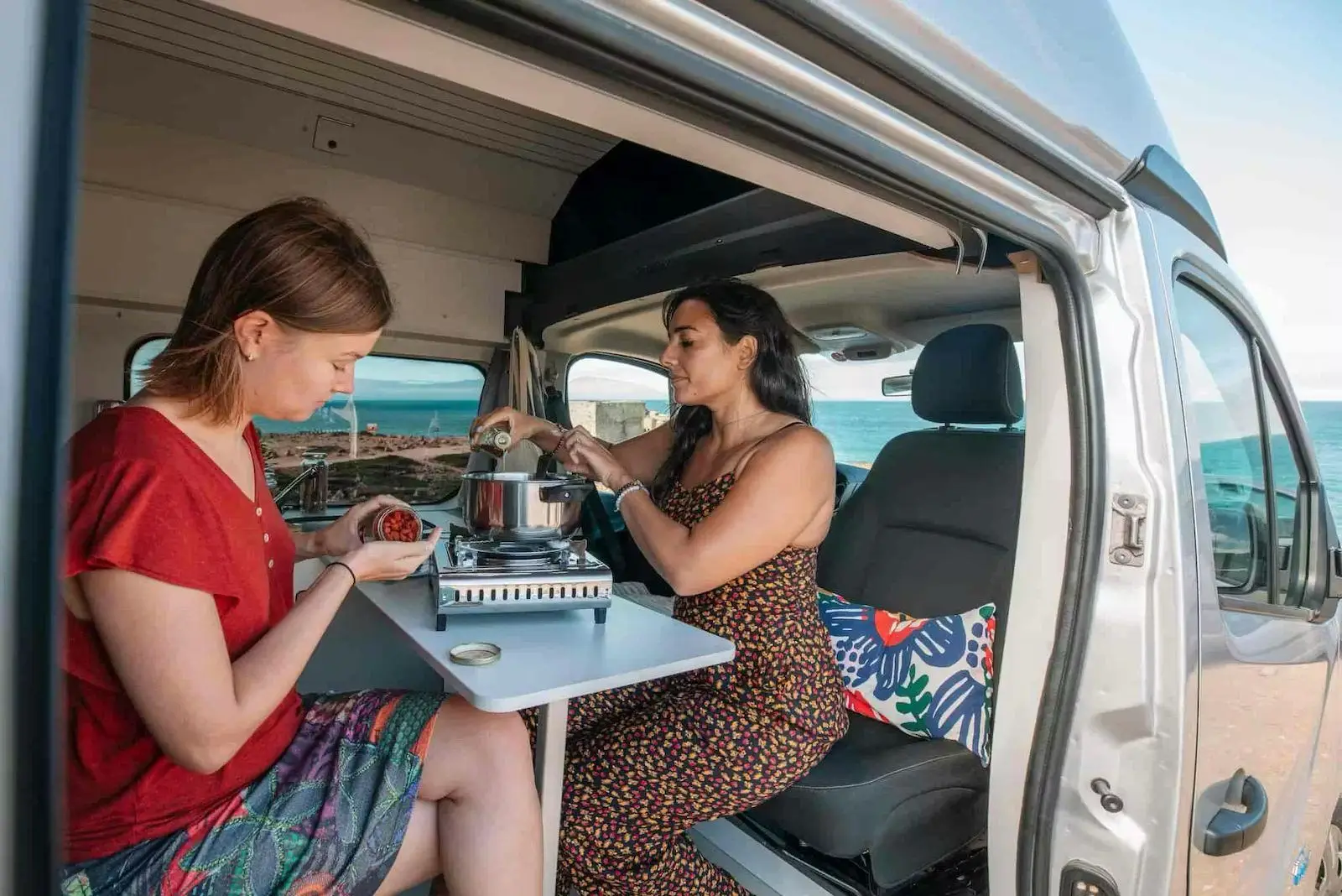 Two people cooking at the table of the Fiat Talento campervan, side door open to the sea